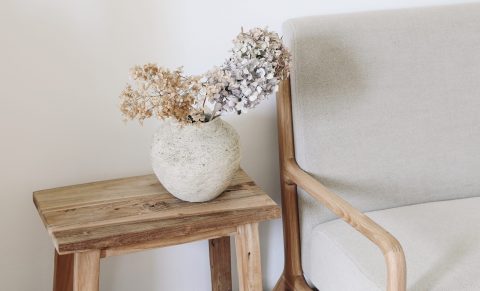 Fall still life photo. Rustic textured vase with dry hydrangea flowers on old wooden bench. Blurred linen mid century sofa background. White wall. Scandinavian interior. Boho elegant home decor.